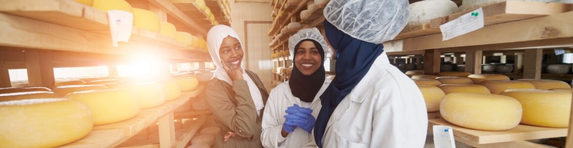 Three women in a room full of cheese in white coats and hair nets helping the cheese production.