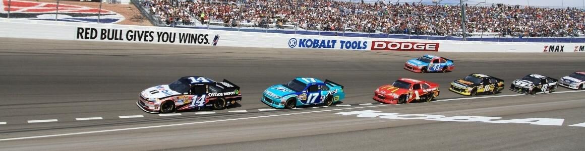 A racetrack with NASCAR drivers racing against each other.